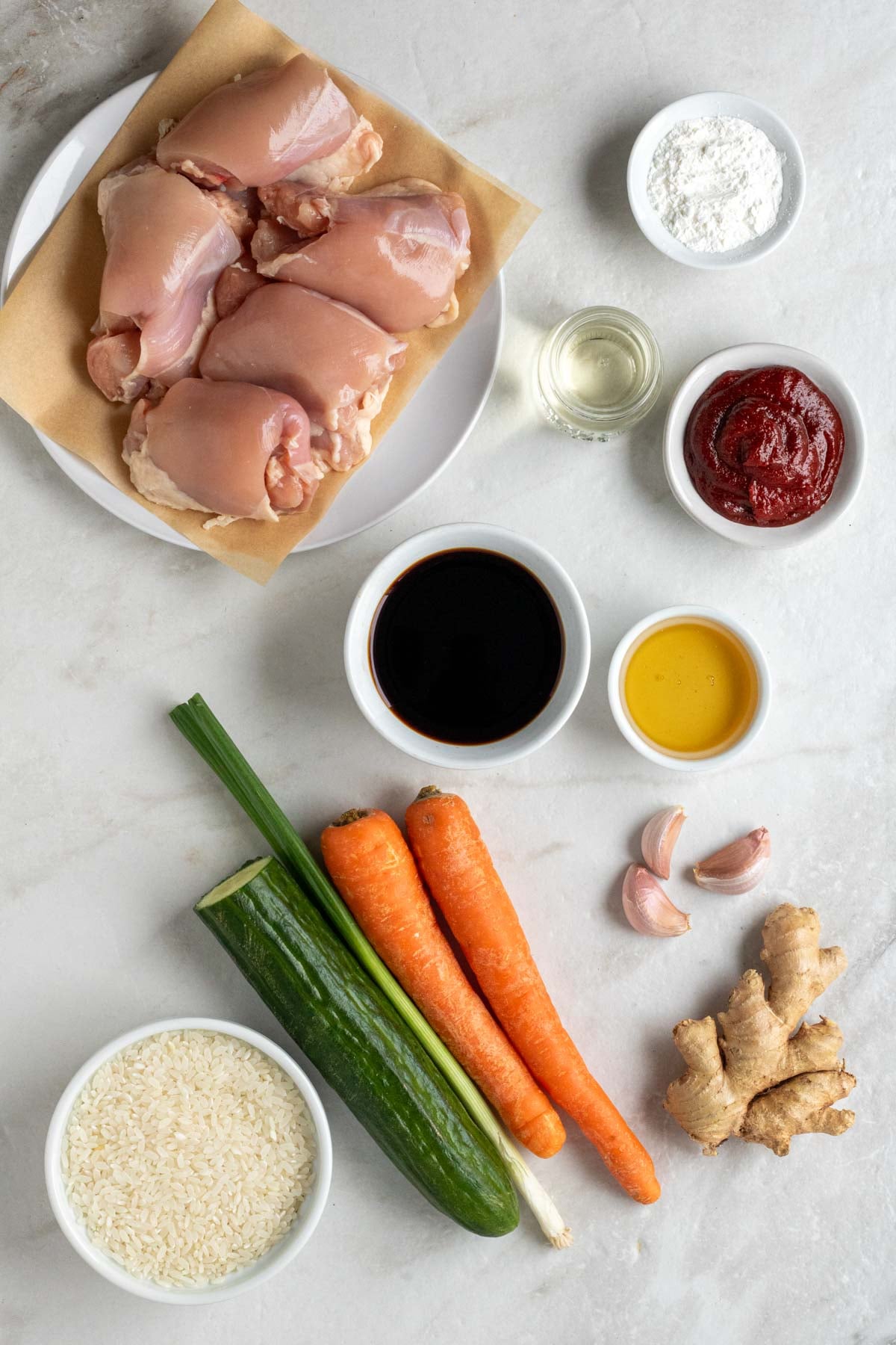 Plate of raw chicken thighs, bowl of corn starch, bowl of rice vinegar, bowl of gochujang paste, bowl of soy sauce, bowl of honey, garlic cloves, ginger root, carrots, green onion, cucumber, and a bowl of white rice on a white background.