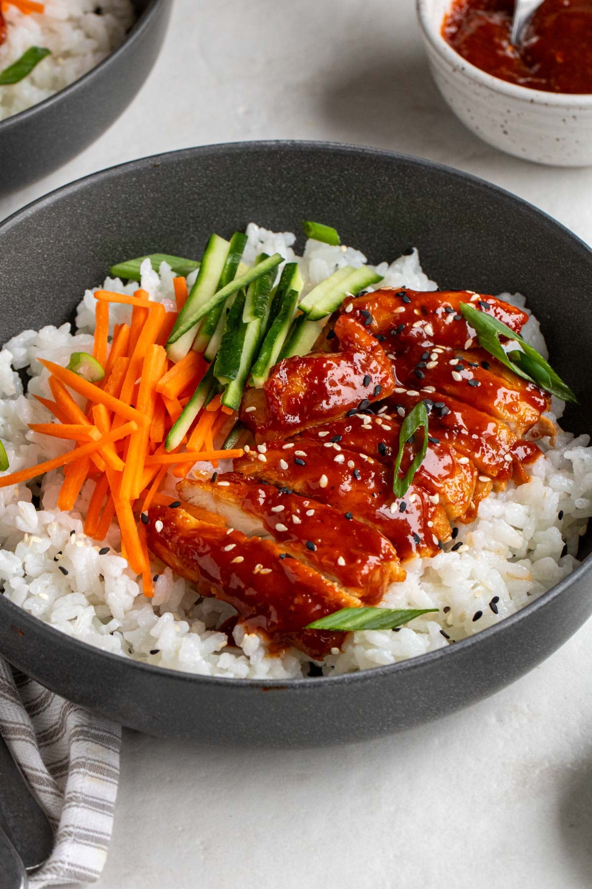 Close up of gochujang chicken thighs on a bed of white rice with sliced carrots, cucumber, green onion, and sesame seeds in a dark grey bowl on a white background.