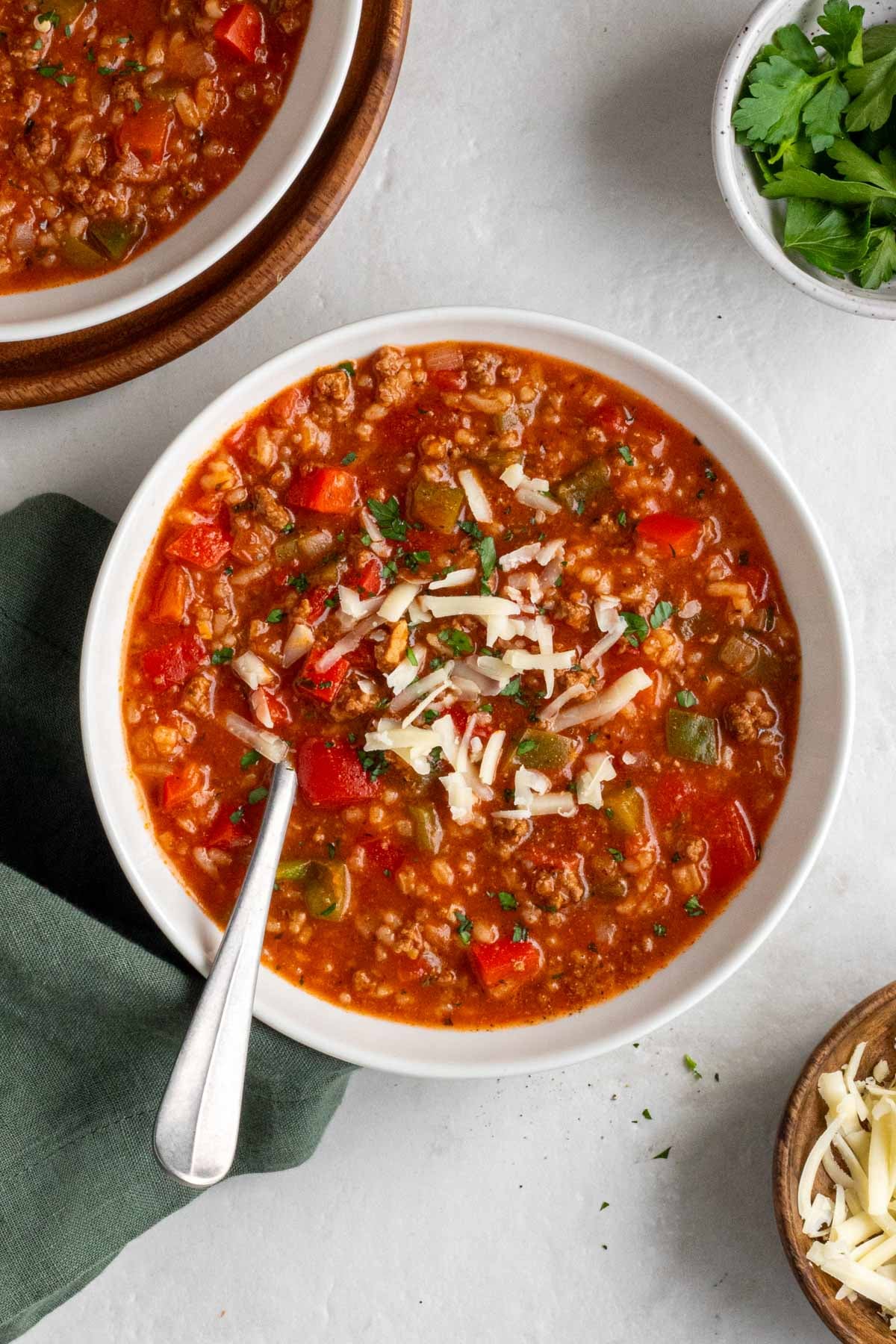Two bowls of stuffed pepper soup with shredded cheese and freshly parsley on top with a spoon and a grey tea towel on a white background.