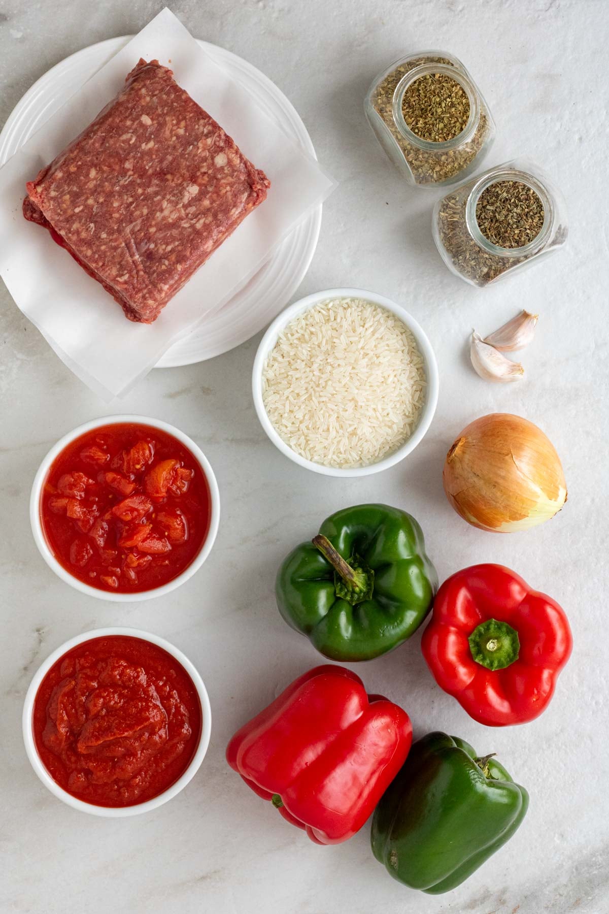Plate of lean ground beef with jars of dried oregano and basil, a bowl of raw white raws, bowls of diced tomatoes and crushed tomatoes, with garlic cloves, onion, and bell peppers on a white background.