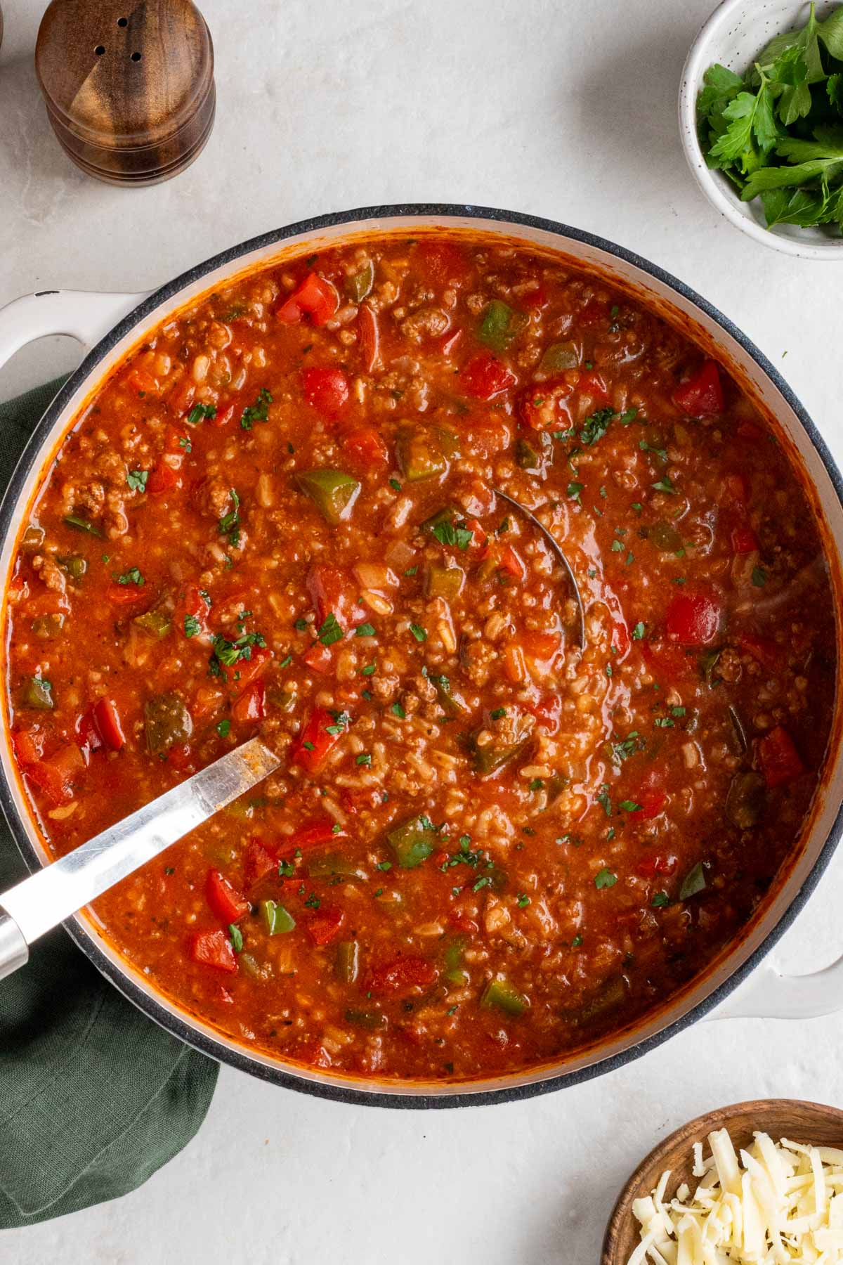 Large pot filled with unstuffed pepper soup with a ladle with a green tea towel, bowl of fresh parsley, and bowl of shredded cheese on the side on a white background.