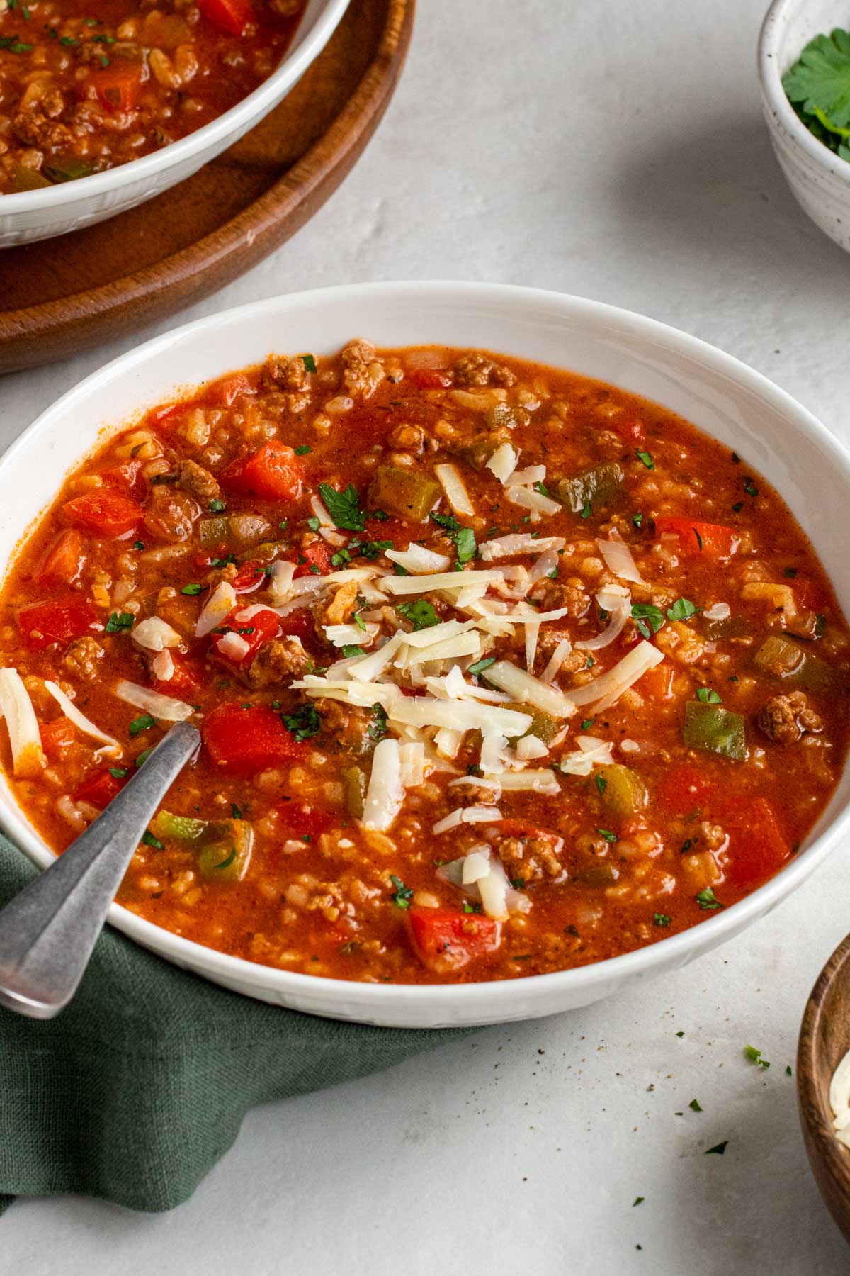 Close up of a bowl of unstuffed pepper soup with a silver spoon and shredded cheese on top on a dark green tea towel on a white background.