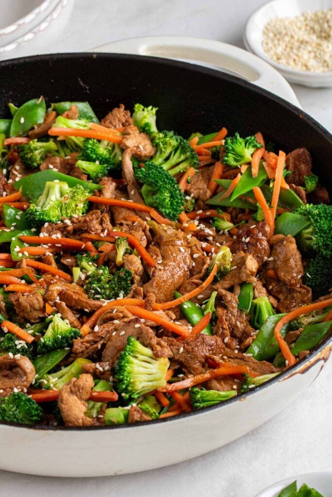 Large pan of ginger pork stir fry with broccoli, carrots, and snow peas with sesame seeds sprinkled on top.
