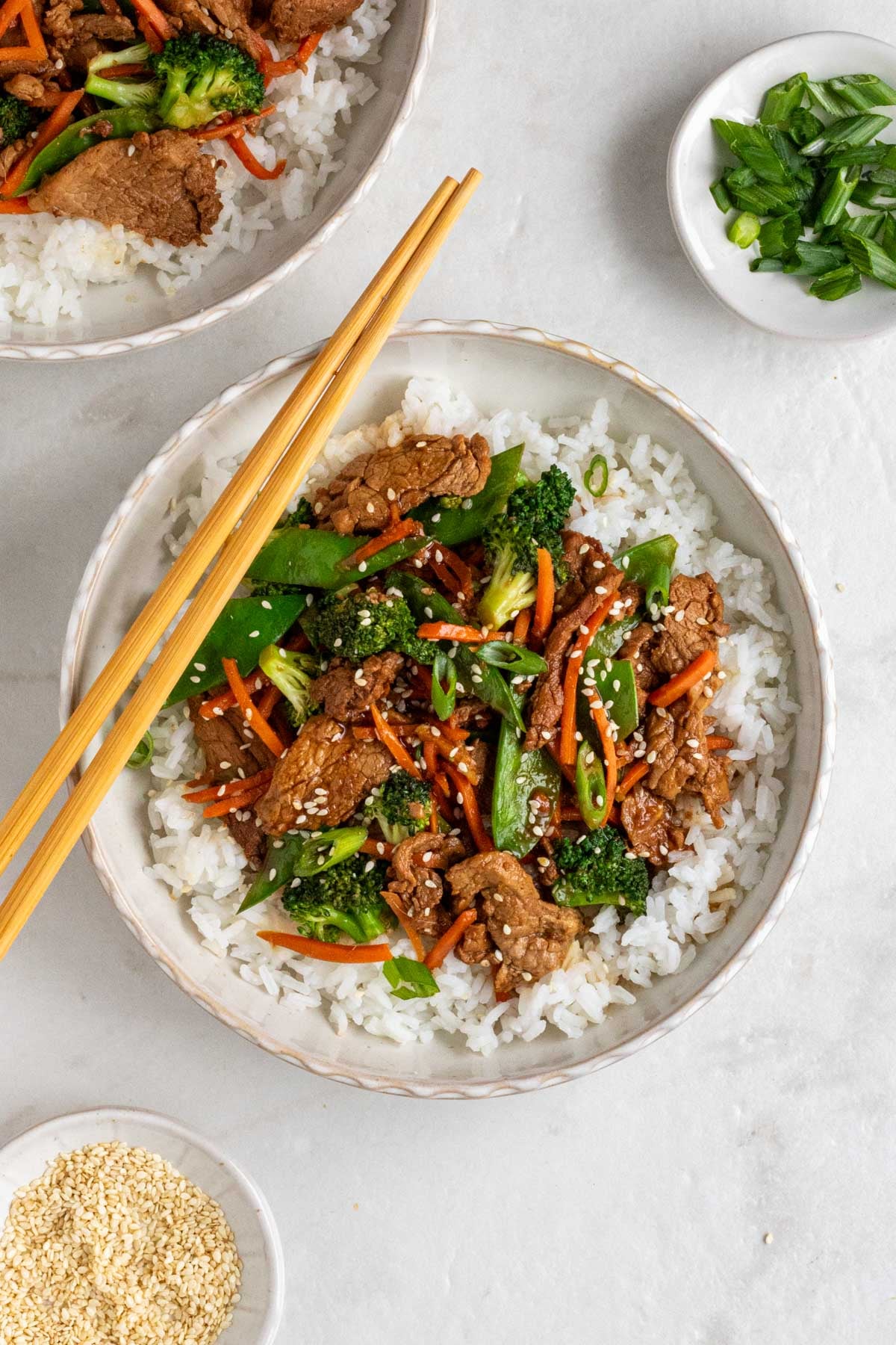 Two white bowls of ginger pork stir fry with white rice with wood chopsticks a side of sesame seeds and sliced green onion on a white background.