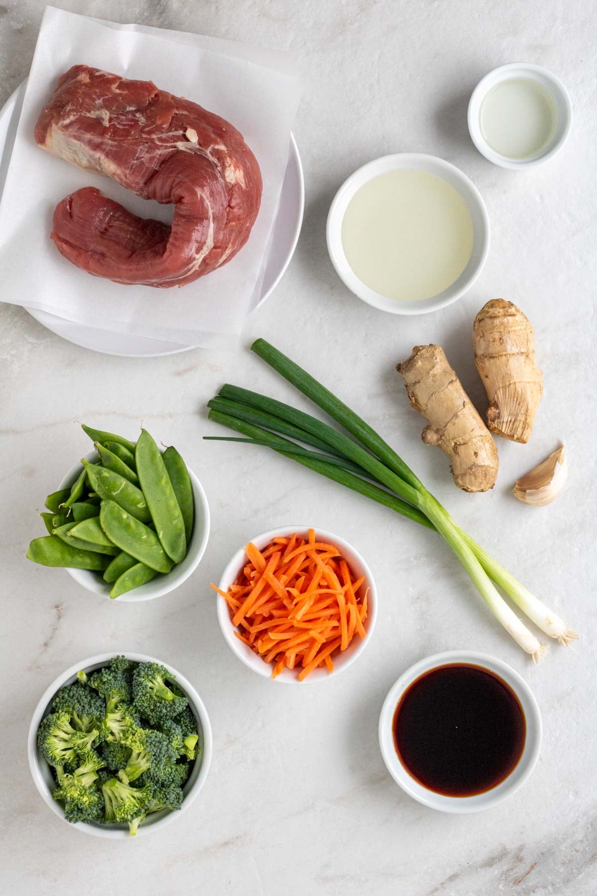 Small plate with pork tenderloin with small white bowls of rice vinegar, mirin, soy sauce, snow peas, broccoli, shredded carrots, green onions, fresh ginger, and garlic cloves on a white background.