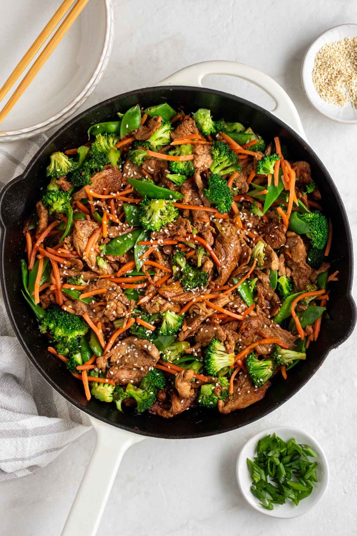 Large cast-iron skillet of ginger pork stir-fry with on a white background with two bowls, wood chopsticks, sesame seeds, and sliced green onion on the side. 