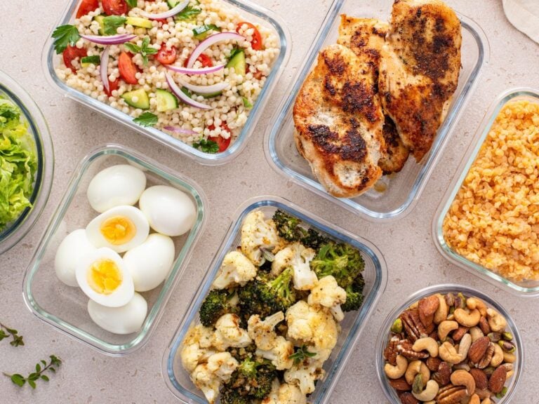 Glass meal prep containers of couscous salad, roasted chicken, cooked lentils, lettuce, hard-boiled eggs, roasted veggies, and nuts on a light pink background.