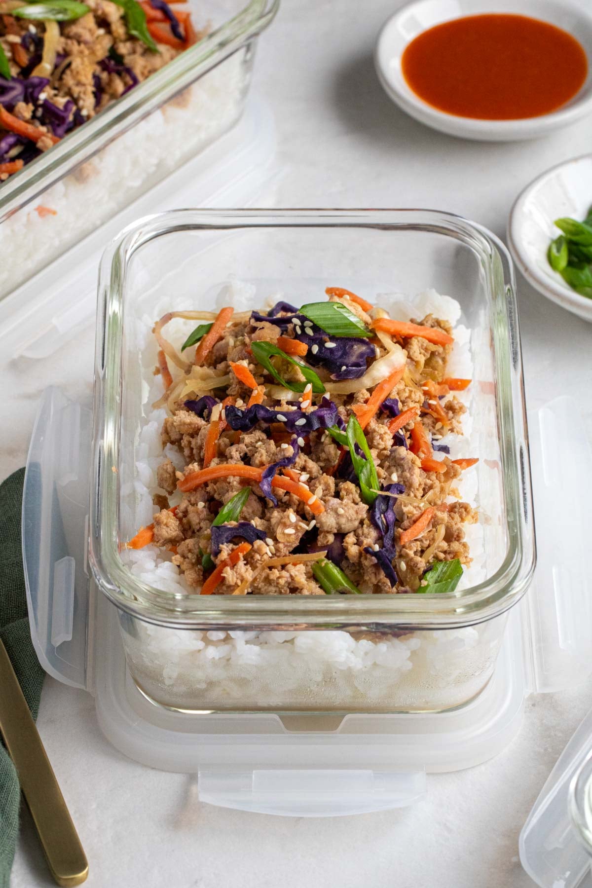 Close up of a deconstructed egg roll in a bowl in a glass meal prep container with a small bowl of hot sauce and sliced green onion in the background and a gold fork and green tea towel on the side.