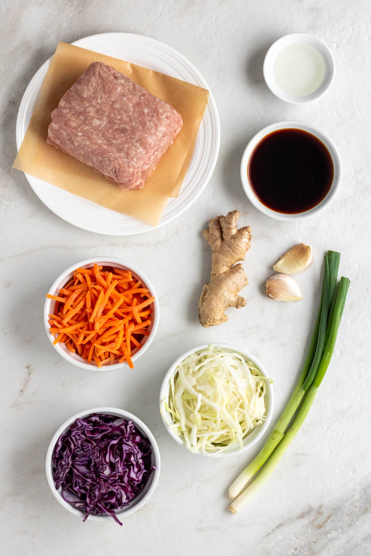Plate of ground pork, small bowl of rice vinegar, small bowl of soy sauce, fresh ginger, garlic cloves, green onions, and bowls of shredded carrots, green cabbage, and red cabbage on a white background.