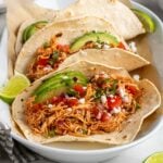 White platter filled with slow cooker chicken tacos topped with sliced avocado, salsa, red onion, crumbled feta cheese, and chopped cilantro with lime wedges on the side on a white background.