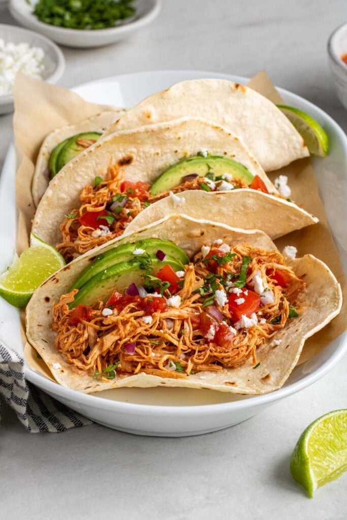White platter filled with slow cooker chicken tacos topped with sliced avocado, salsa, red onion, crumbled feta cheese, and chopped cilantro with lime wedges on the side on a white background.