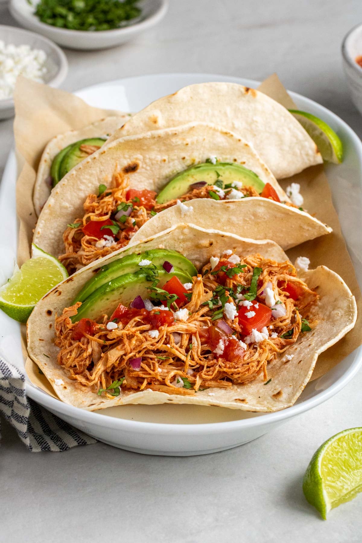 White platter filled with slow cooker chicken tacos topped with sliced avocado, salsa, red onion, crumbled feta cheese, and chopped cilantro with lime wedges on the side on a white background.
