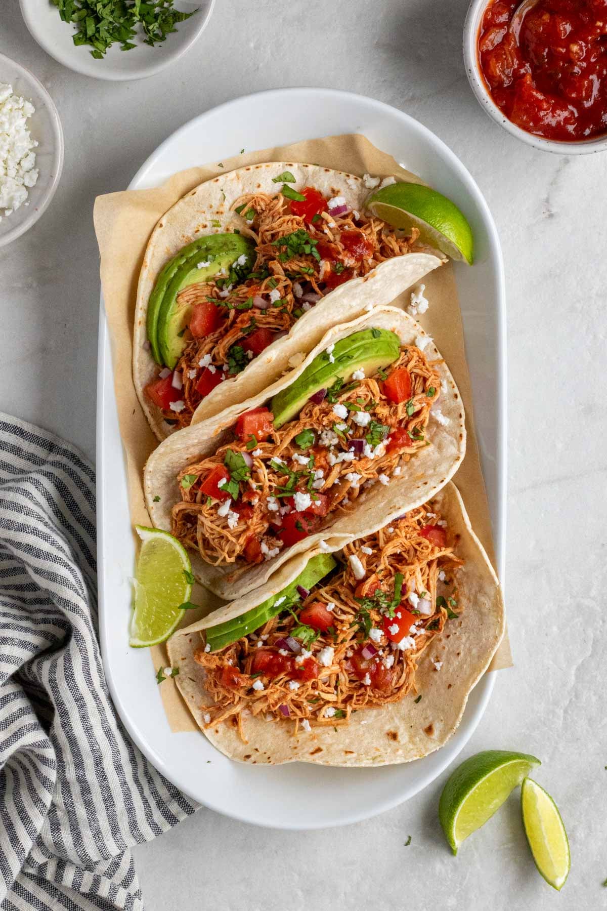 Overhead image of a plate of three crockpot shredded chicken tacos with feta, cilantro, salsa, and lime wedges on the side with a striped blue and white tea towel on a white background.