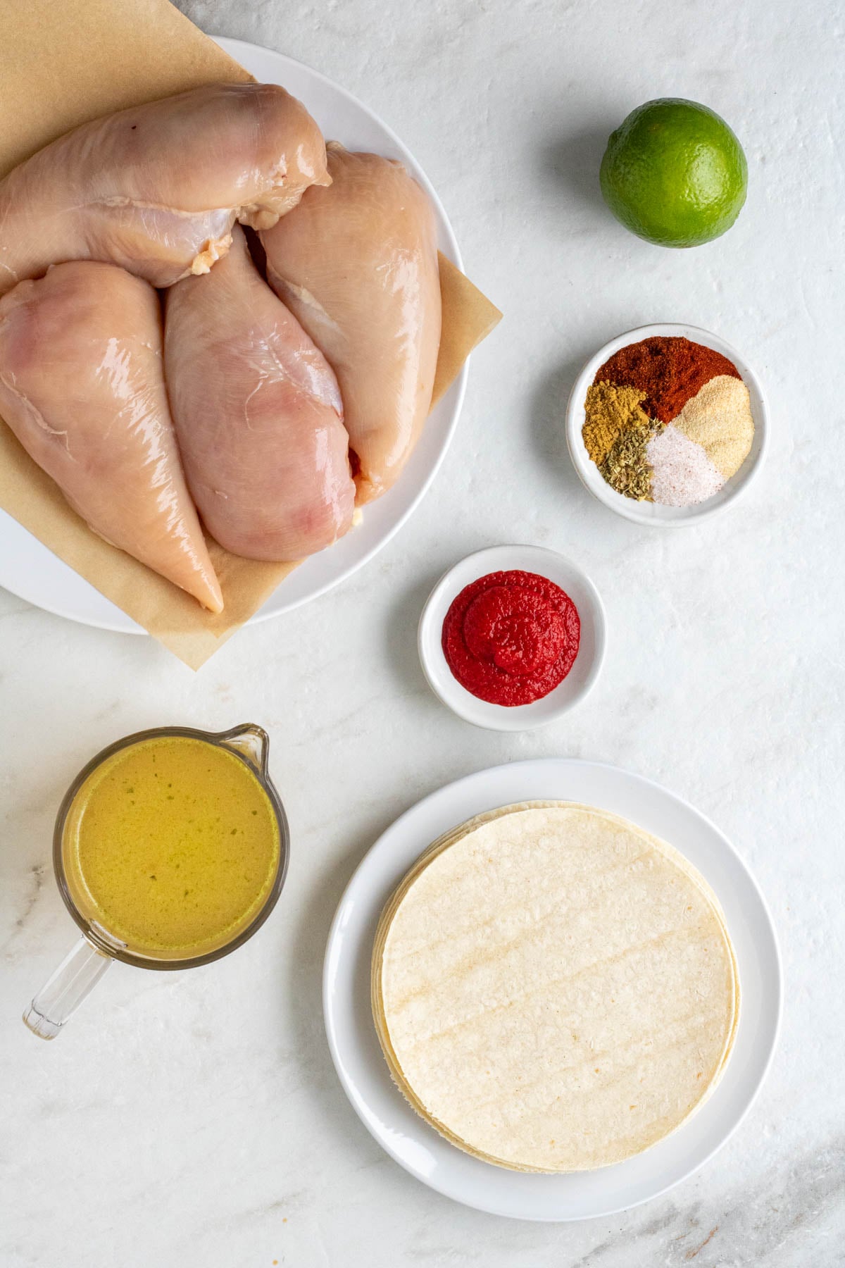 Plate of raw chicken breasts, lime, small bowl of spices, small bowl of tomato paste, jug of chicken broth, and a plate of corn tortillas on a white background.