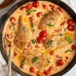 Baked feta chicken is a large cast-iron skillet with fresh basil on top with a gold serving spoon and a beige checkered tea towel on the side on a white background.