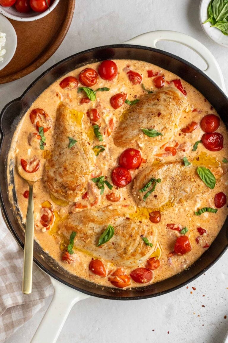 Baked feta chicken is a large cast-iron skillet with fresh basil on top with a gold serving spoon and a beige checkered tea towel on the side on a white background.