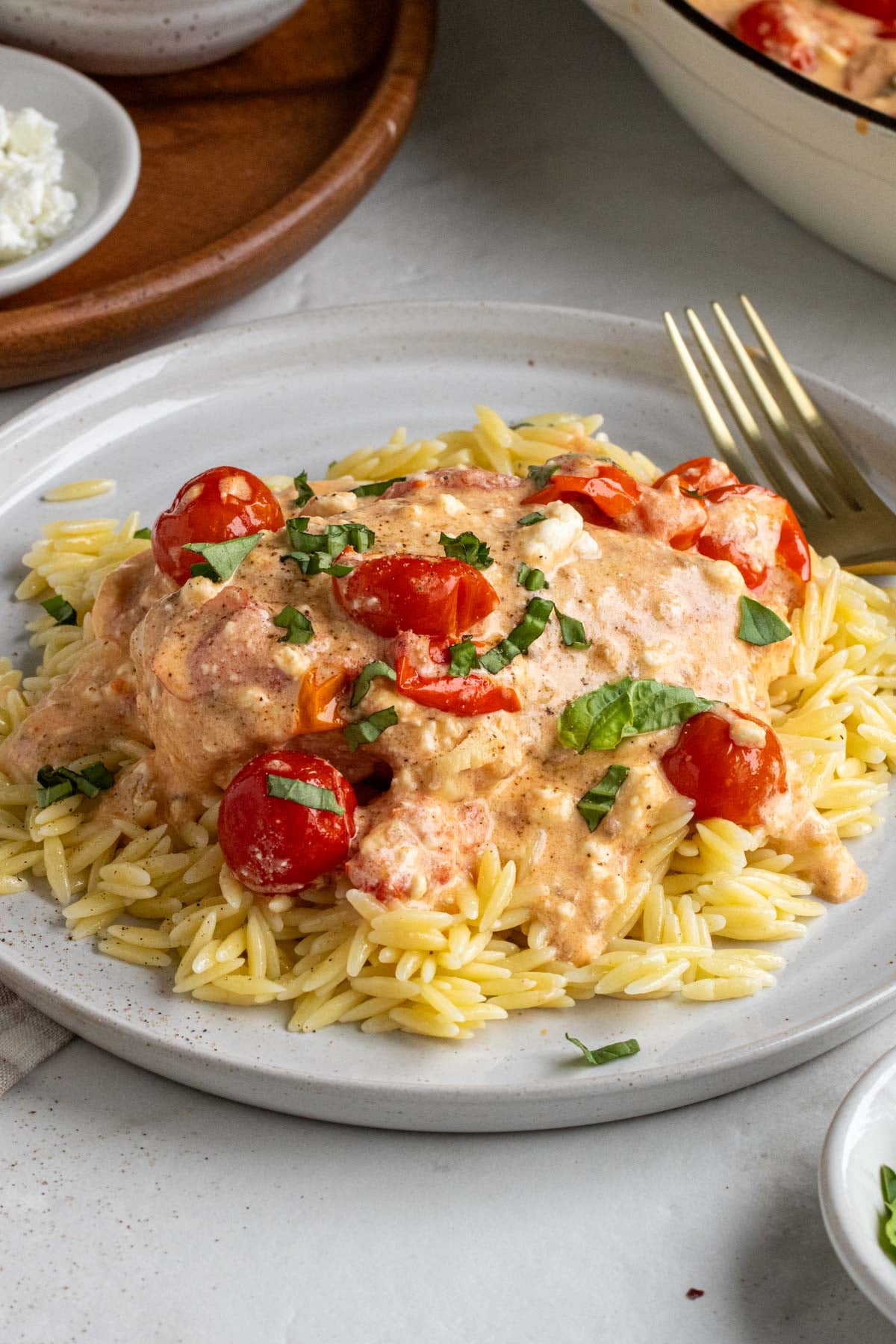 Small beige plate with baked feta chicken on top of cooked orzo with fresh basil on top with a gold fork on a white background.