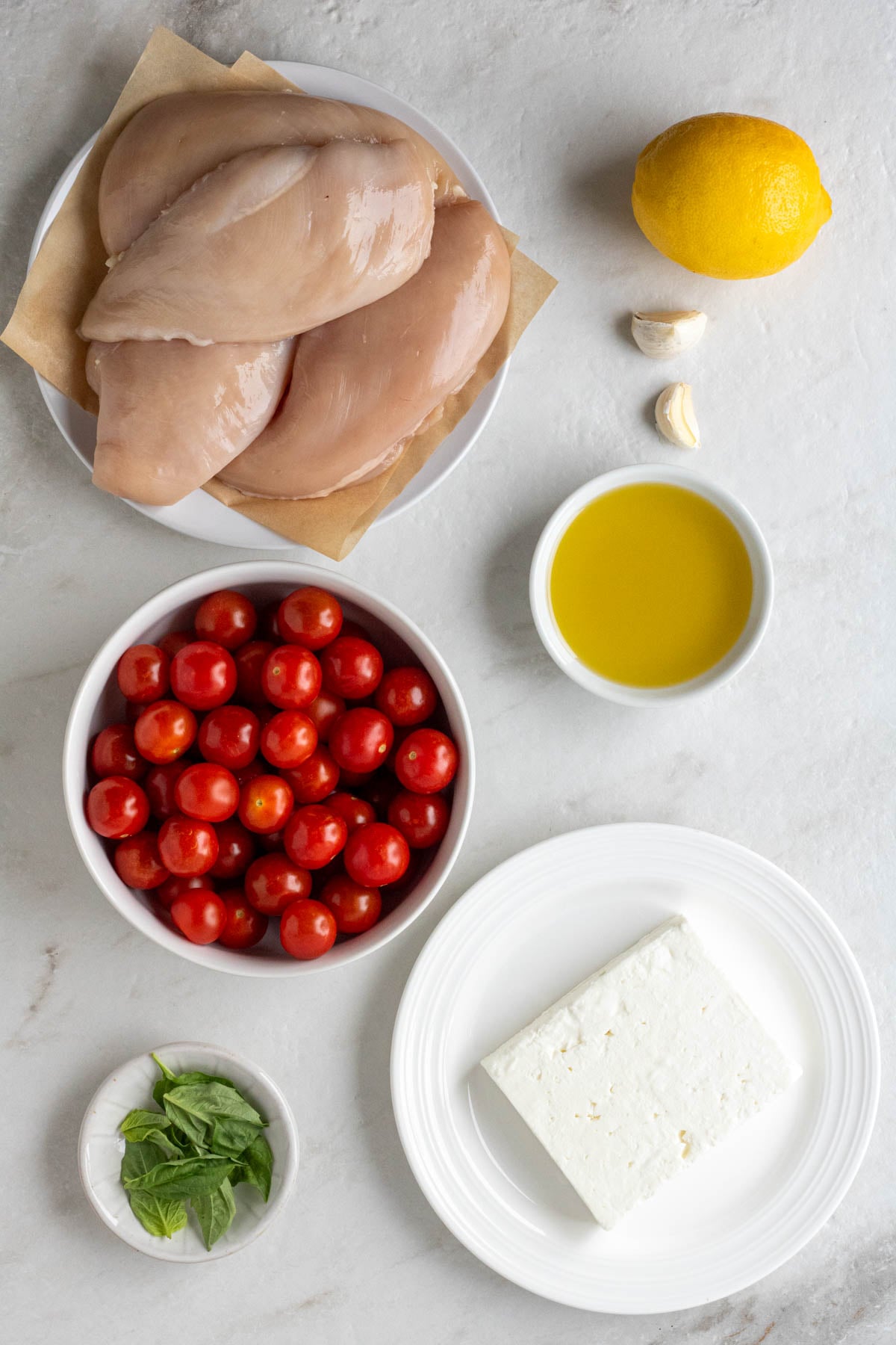 White plate with raw chicken breasts, lemon, garlic cloves, bowl of olive oil, bowl of cherry tomatoes, bowl of basil leaves, and a plate with a block of feta cheese on a white background.