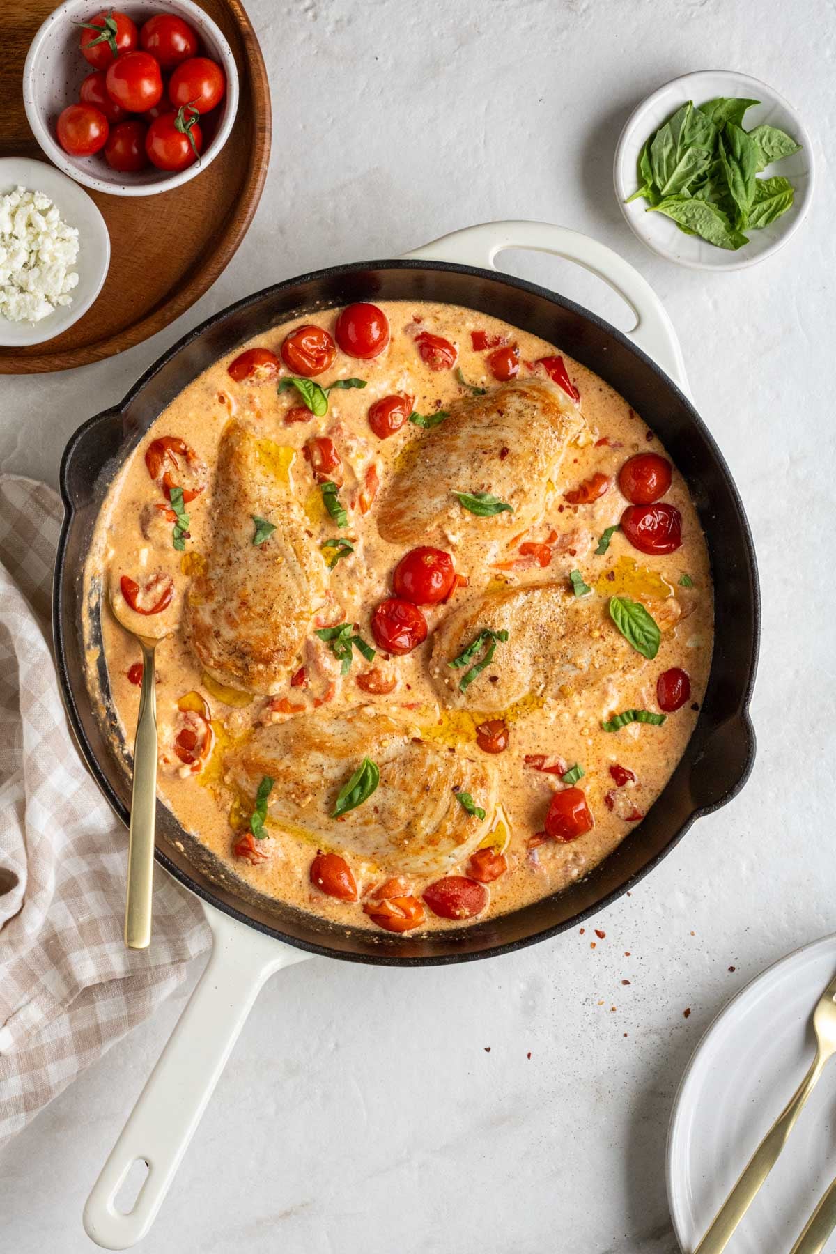 Large cast-iron skillet filled with feta baked chicken with fresh basil on top with a gold serving spoon with a bowl of ripe cherry tomatoes, crumbled feta cheese, and fresh basil leaves on the side with a checkered beige tea towel on a white background.