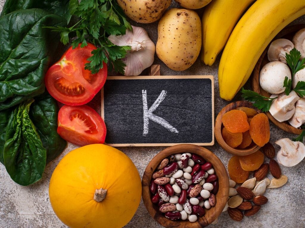 A small chalkboard with the letter K on surrounded by leafy greens, tomatoes, garlic, potatoes, bananas, mushrooms, apricots, almonds, beans, and squash on a dark background.