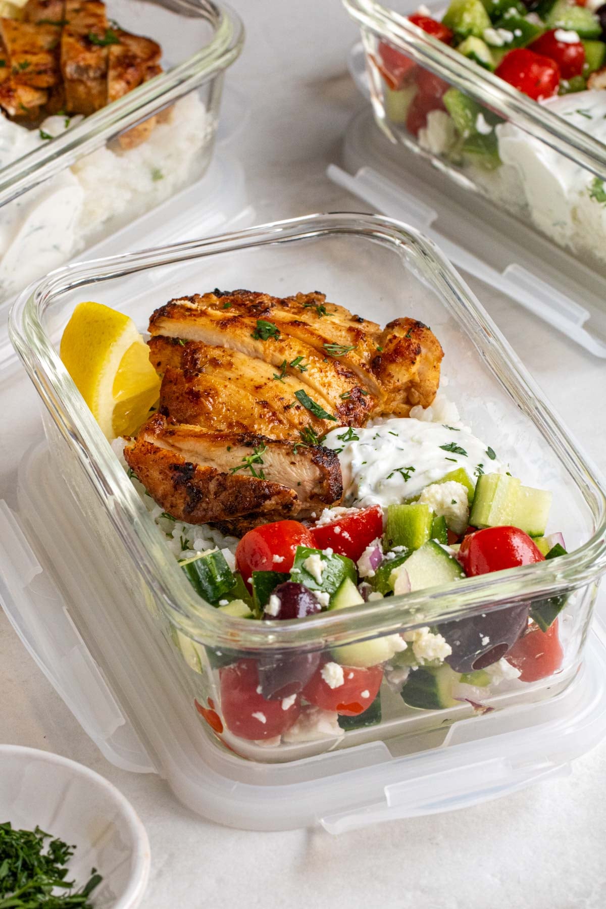 Close up of a Greek chicken bowl with cucumber tomato salad, rice pilaf, and tzatziki in a glass meal prep container.
