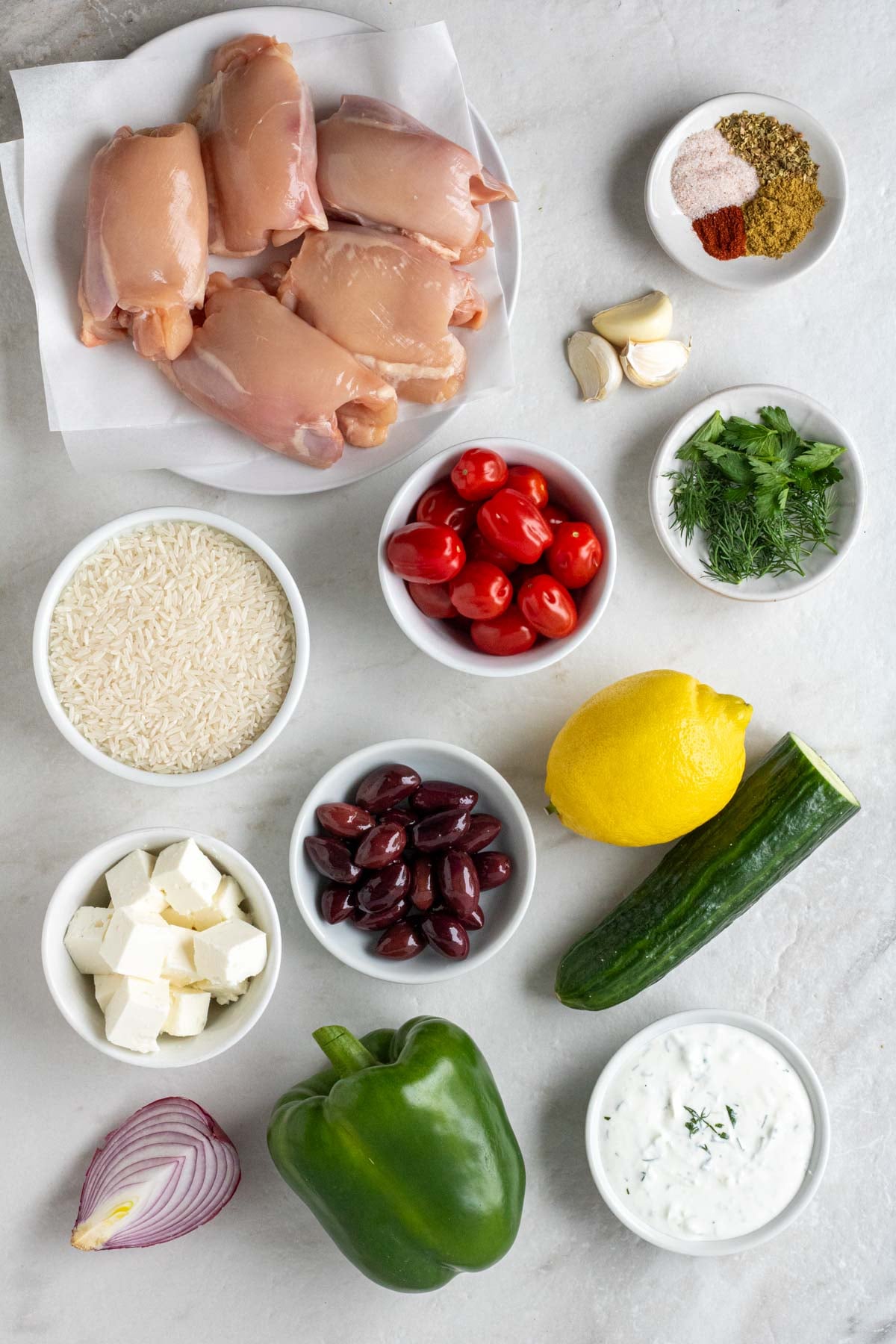 Plate of raw chicken thighs with bowls of spices, fresh herbs, grape tomatoes, rice, olive, feta cheese and tzatziki with garlic cloves, lemon, cucumber, green bell pepper, and red onion on a white background.