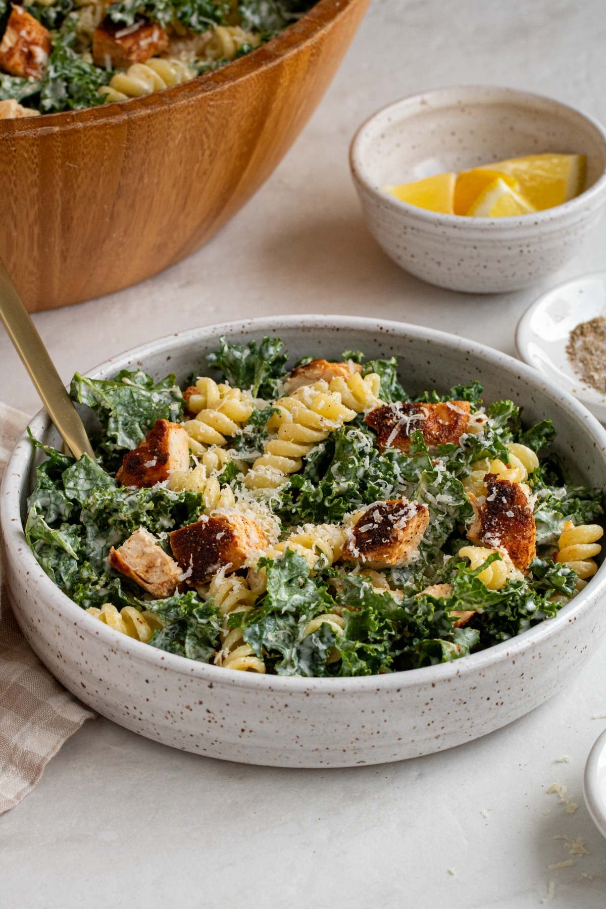 A serving of kale caesar pasta salad with grilled chicken in a beige speckled bowl with a gold fork with a small bowl of lemon sliced and black pepper on the side on a white background.