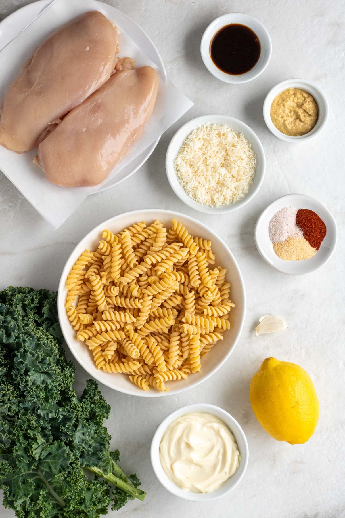 Plate of raw chicken breasts with a bowl of Worcestershire sauce, Dijon mustard, parmesan cheese, spices, fusilli pasta, and mayonnaise with a garlic clove, lemon, and fresh kale on the side on a white background.