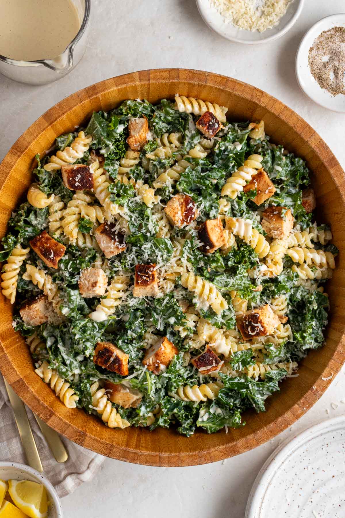 Large wood bowl of kale chicken caesar pasta salad with freshly grated parmesan cheese on top with a glass jug of Caesar dressing, a small bowl of grated parmesan, a small bowl of black pepper, with a beige checkered tea towel with a gold fork and spoon on the side.