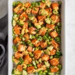 Salmon sushi bake in a large white casserole dish topped with spicy mayo and furikake and bowls of sliced green onion and sesame seeds on the side with a grey tea towel on a white background.