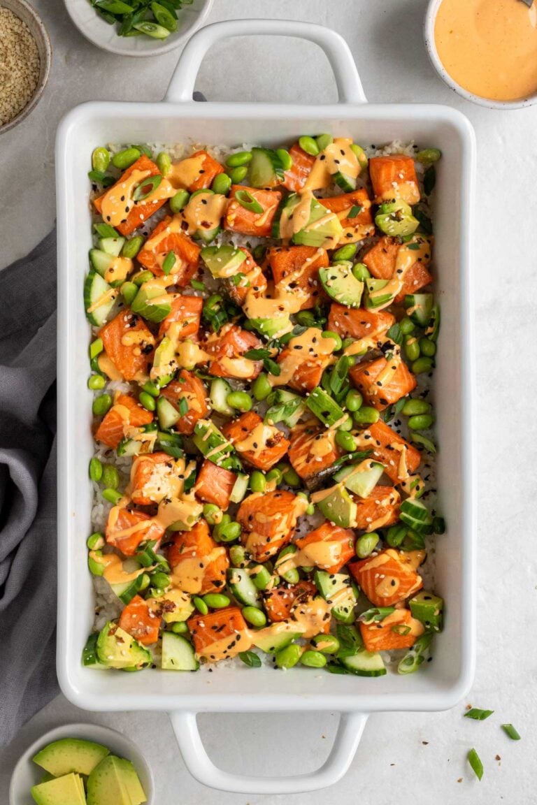 Salmon sushi bake in a large white casserole dish topped with spicy mayo and furikake and bowls of sliced green onion and sesame seeds on the side with a grey tea towel on a white background.