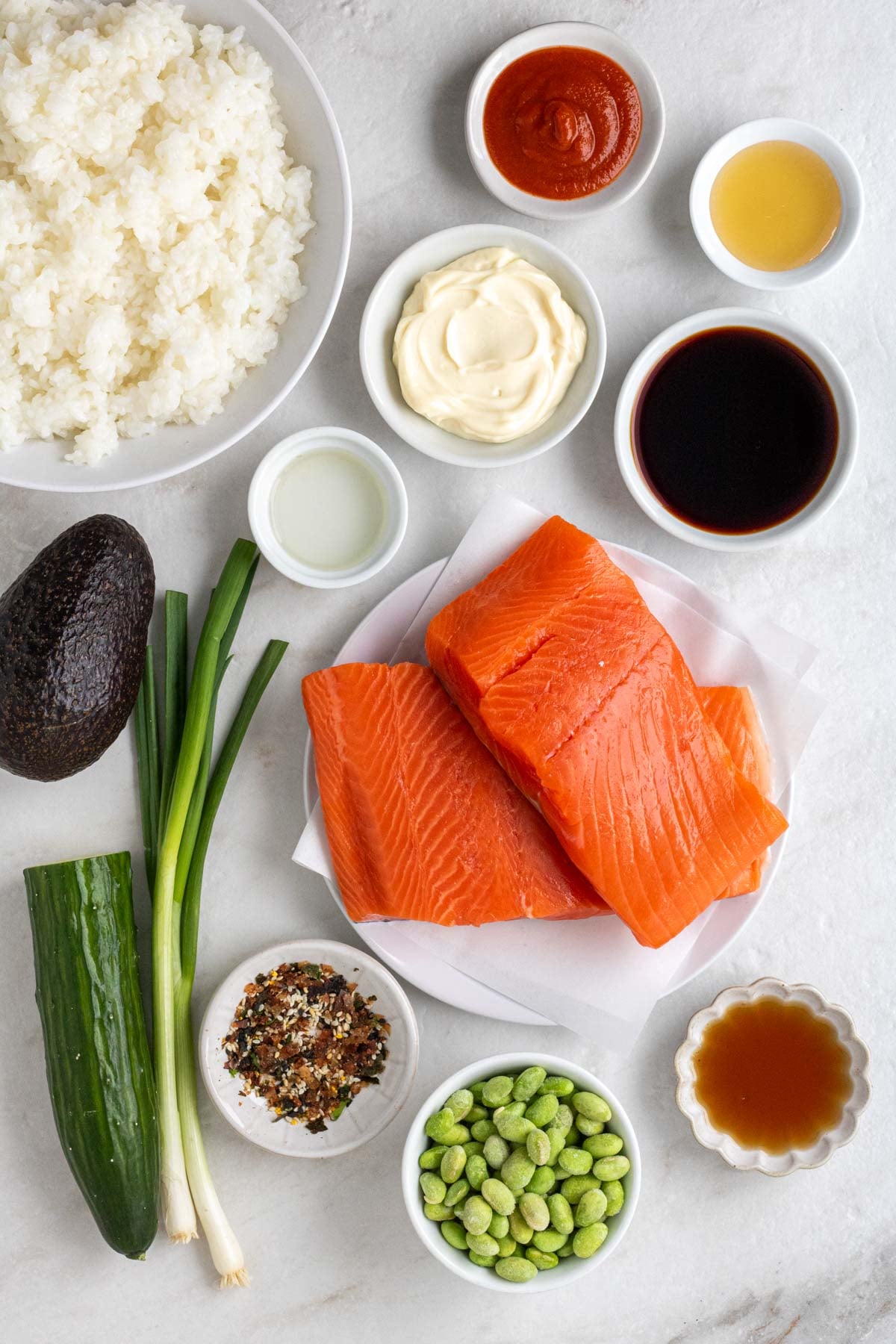 Bowls of cooked sushi rice, sriracha, honey, soy sauce, mayonnaise, rice vinegar, sesame oil, frozen edamame, and furikake with a white plate with raw salmon fillet and a whole avocado, green onions, and half a cucumber on a white background. 
