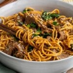 Close up of a beige bowl of sesame beef noodles wrapped around dark wood chopstick with a green tea towel on a white background.
