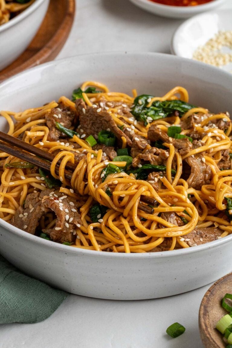 Close up of a beige bowl of sesame beef noodles wrapped around dark wood chopstick with a green tea towel on a white background.