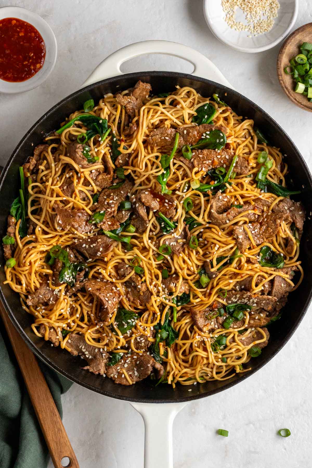 Large pan of sesame beef stir-fry noodles with small bowls chili sauce, sesame seeds, and sliced green onions on the side with a dark green tea towel and a wooden spoon on a wide background.