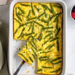 Baking dish of asparagus egg bake with a slice of a silver serving spatula with sliced bread, a bowl of leafy greens, a small bowl of fresh berries, and a small bowl of crumbled feta cheese on the side.