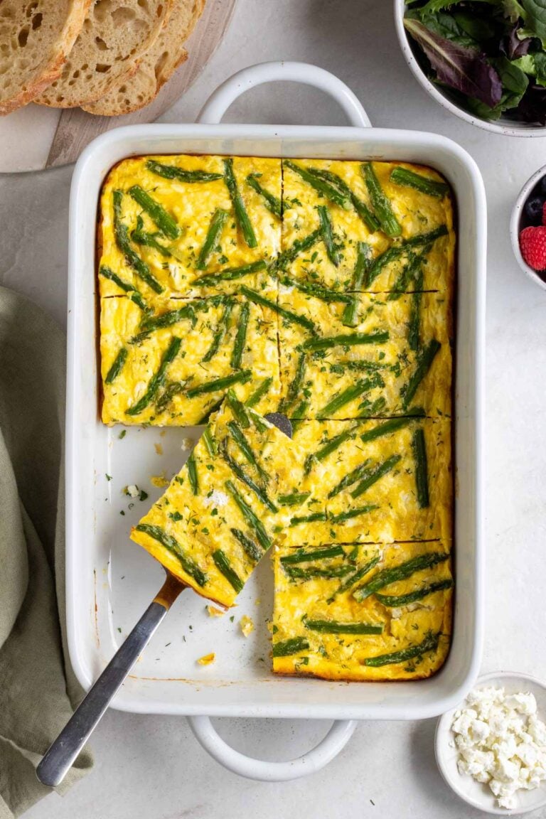 Baking dish of asparagus egg bake with a slice of a silver serving spatula with sliced bread, a bowl of leafy greens, a small bowl of fresh berries, and a small bowl of crumbled feta cheese on the side.
