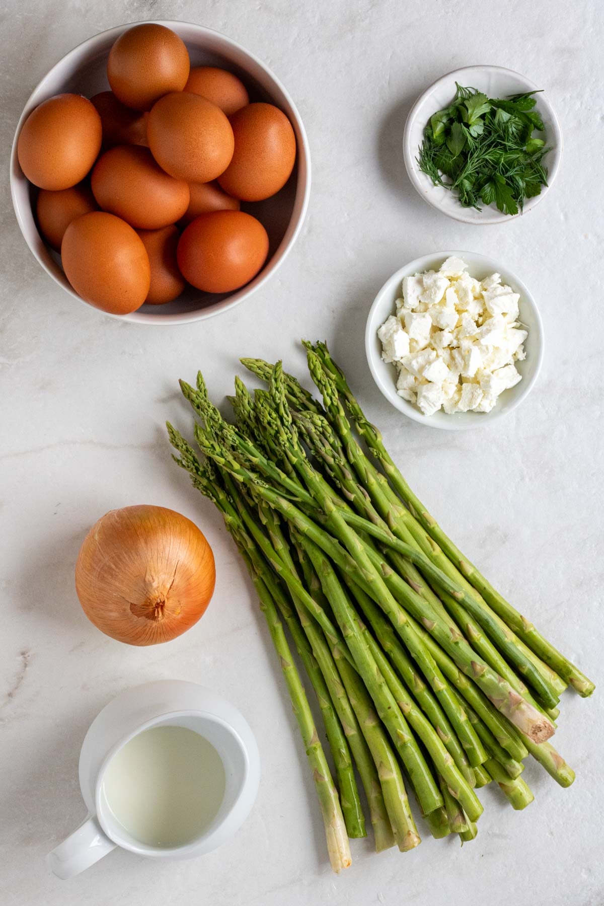 Bowls of eggs, fresh herbs, and crumbled feta cheese with a bunch of fresh, raw asparagus, an onion, and a jug of milk on a white background.