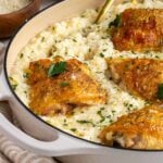 Large beige enabled cast-iron pan with chicken and leek risotto on a beige and striped tea towel with a gold serving spoon and a small wood bowl of freshly grated parmesan cheese and small white bowl of fresh parsley in the background.
