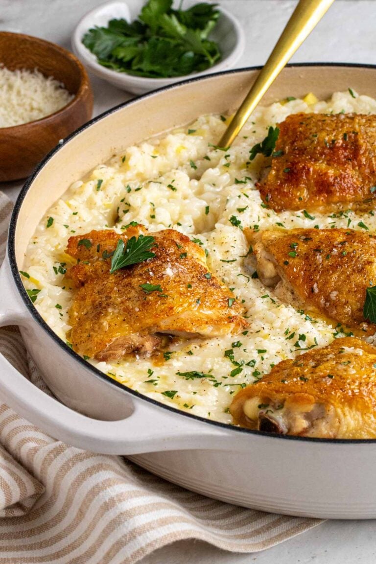 Large beige enabled cast-iron pan with chicken and leek risotto on a beige and striped tea towel with a gold serving spoon and a small wood bowl of freshly grated parmesan cheese and small white bowl of fresh parsley in the background.