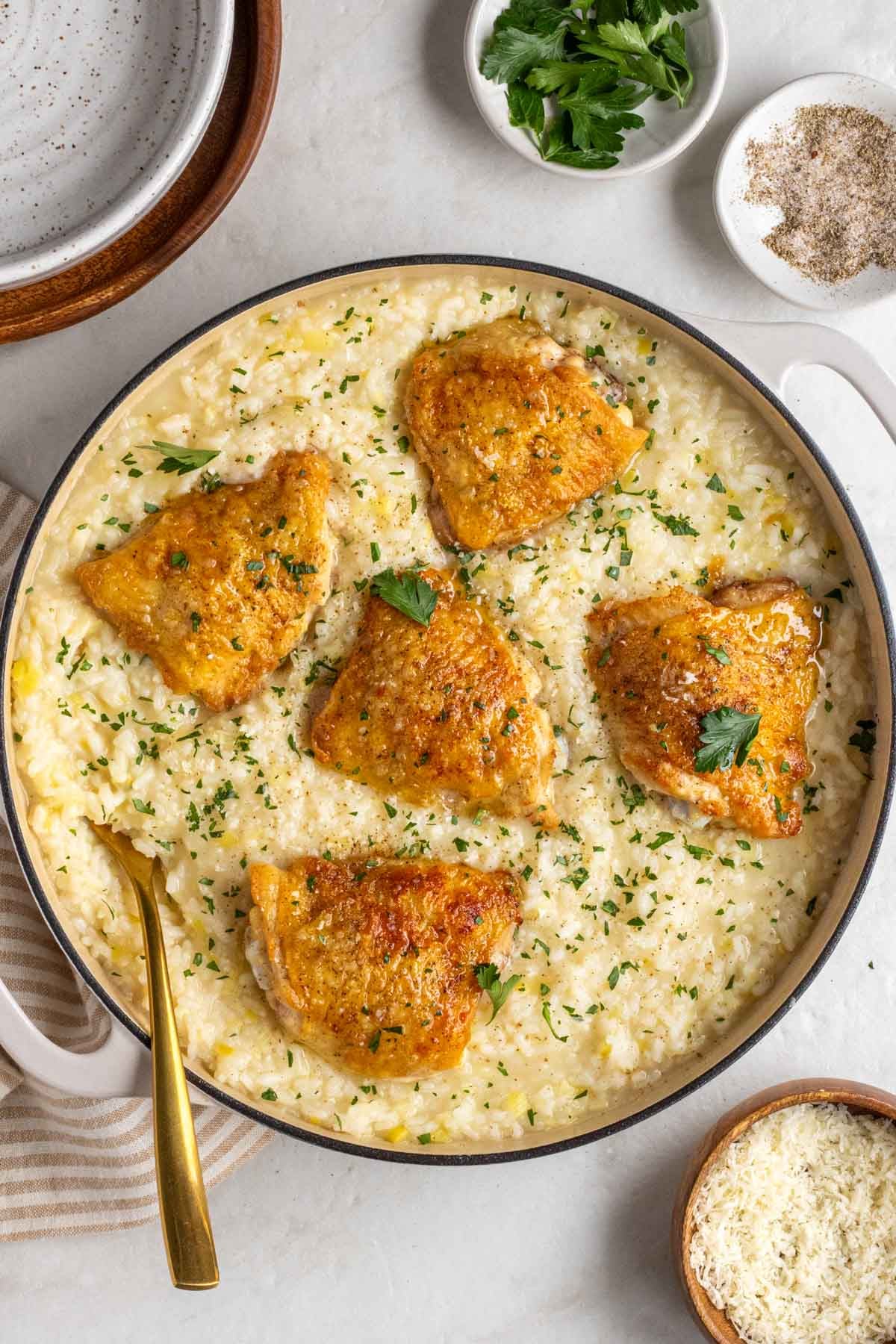 Large pot of leek risotto with chicken thighs on top with a gold serving spoon on a beige tea towel with parmesan cheese and parsley on the side on a white background.