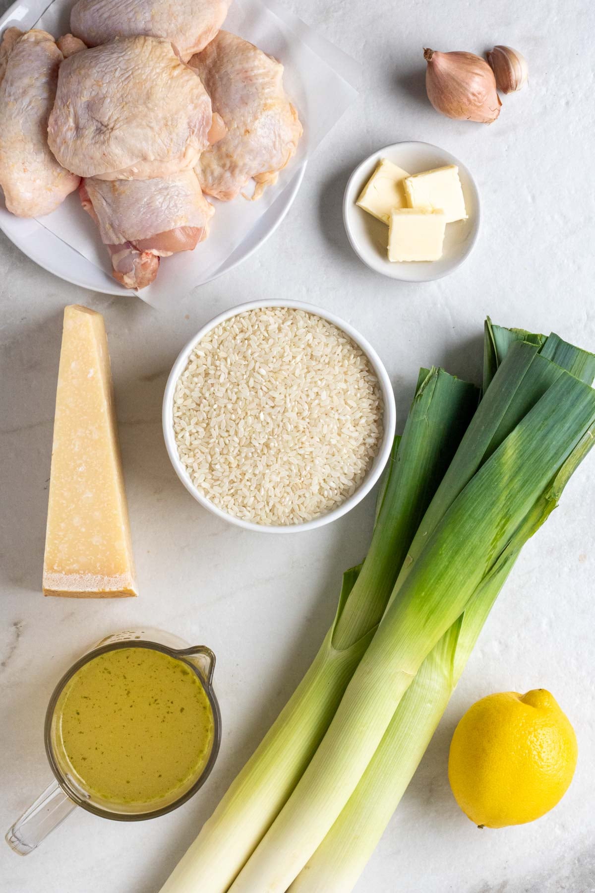 Plate of raw chicken thighs, shallot, garlic cloves, bowl of butter, bowl of arborio rice, parmesan cheese, glass jug of chicken stock, leeks, and a lemon on a white background.