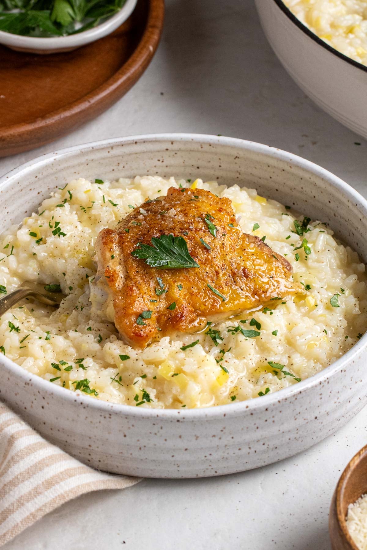 White speckled bowl filled with a serving leek risotto with a chicken thigh on top with a gold fork on a white background.