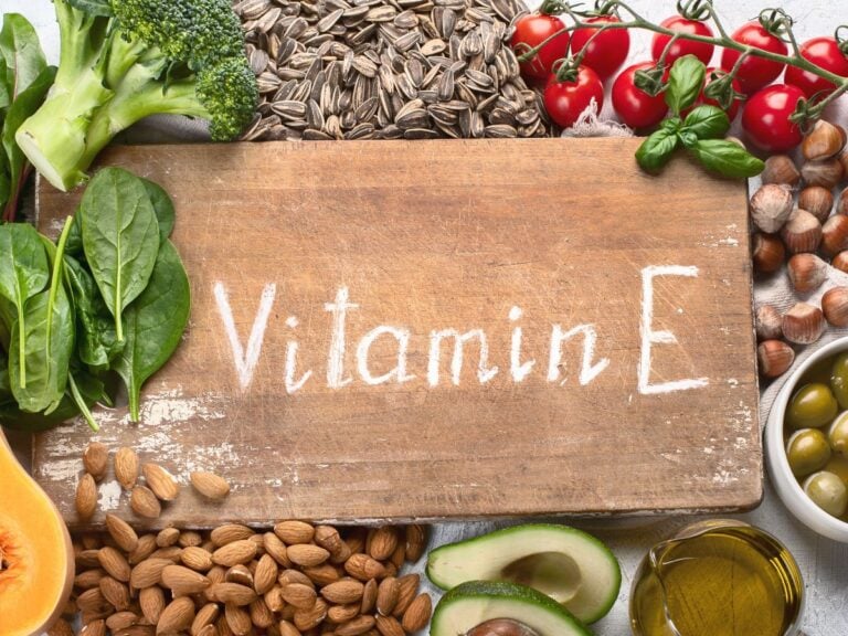 Wood cutting board with the words vitamin E written on it in chalk surrounded by broccoli florets, sunflower seeds, cherry tomatoes, hazelnuts, olive, oil, avocado, almonds, and butternut squash.