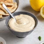 Small grey bowl of garlic tahini sauce with a small spoon with pita bread, lemons, and fresh parsley on the side on a white background.