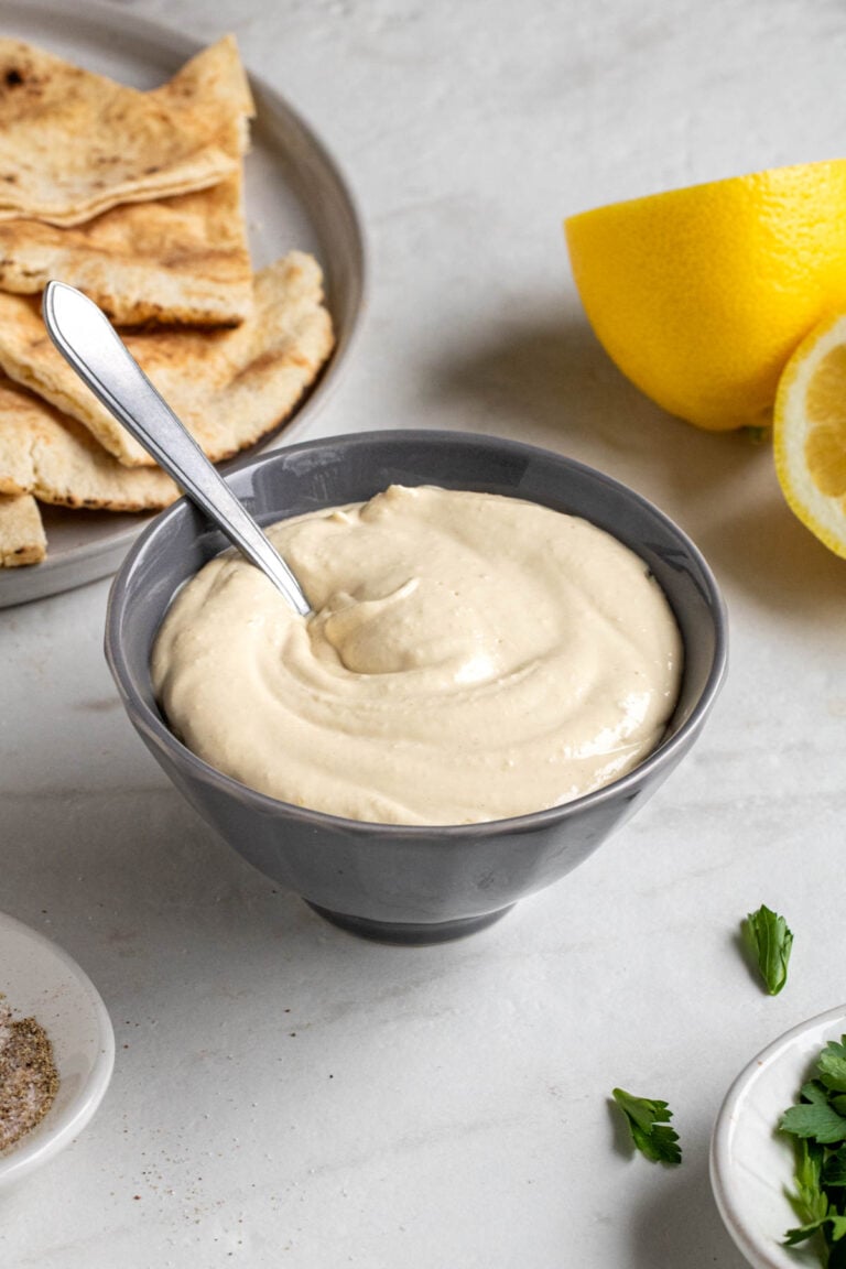 Small grey bowl of garlic tahini sauce with a small spoon with pita bread, lemons, and fresh parsley on the side on a white background.