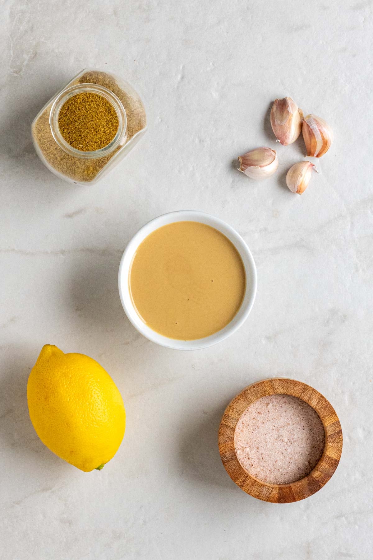 Jar of cumin, garlic cloves, bowl of tahini, lemon, and wood bowl of salt on a white background.