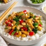 White bowl of honey cashew chicken stir fry with chopsticks and small bowls of sliced green onion, sesame seeds, and cashews on the side.