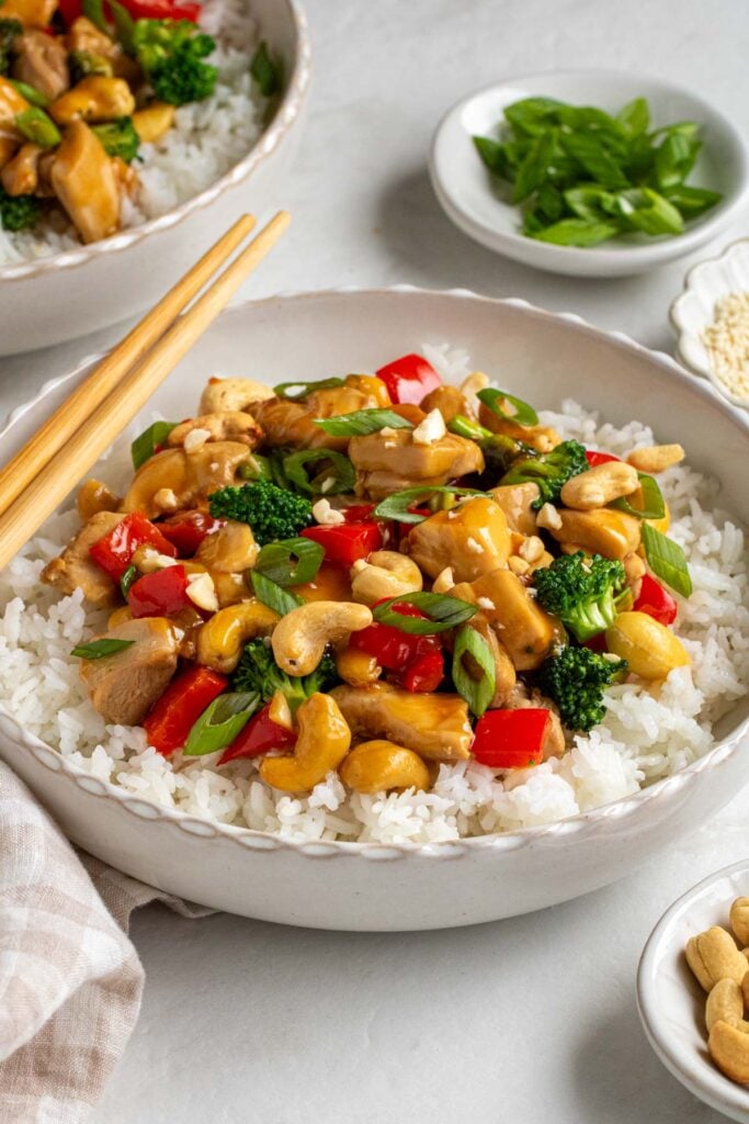 White bowl of honey cashew chicken stir fry with chopsticks and small bowls of sliced green onion, sesame seeds, and cashews on the side.
