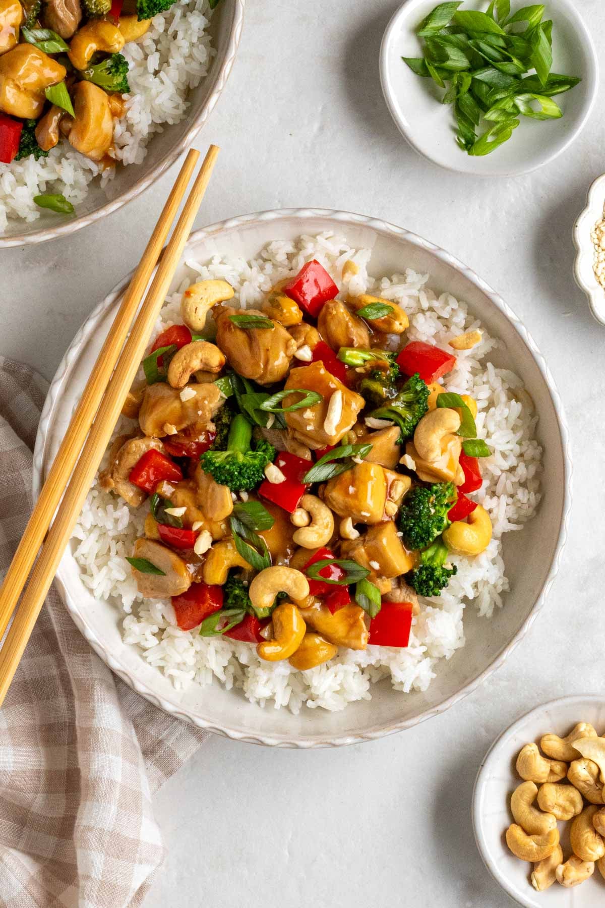 Two bowls honey cashew chicken on top of white rice with sliced green onion and roasted cashews on top with a beige and white checkered tea towel on the side on a white background.